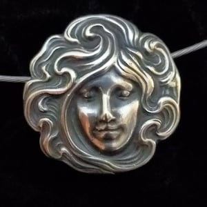 Puede incluir: Un colgante de plata con un rostro estilizado y detallado de una mujer con el pelo suelto. El colgante tiene forma circular y un acabado liso y pulido.