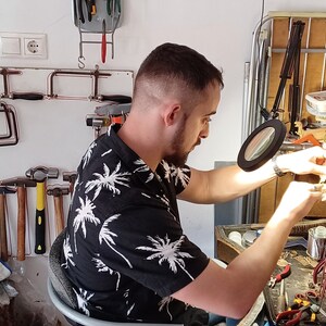 May include: A person working in a workshop, using tools. The person is wearing a black shirt with a white palm tree pattern. Various tools are visible, including hammers, saws, and a magnifying glass, suggesting jewelry making or a similar craft.