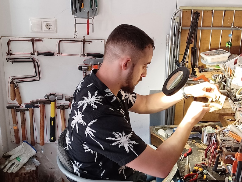 May include: A person working in a workshop, using tools. The person is wearing a black shirt with a white palm tree pattern. Various tools are visible, including hammers, saws, and a magnifying glass, suggesting jewelry making or a similar craft.
