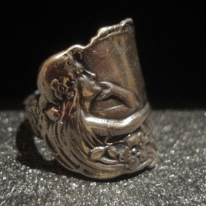 May include: A silver-toned ring with an ornate design. The ring features a sculpted floral and figure motif, with a detailed, textured surface. The ring's design includes a curved, open top, and is set against a dark background.