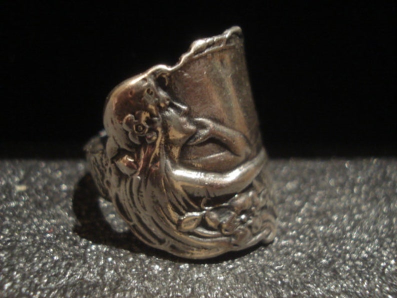 May include: A silver-toned ring with an ornate design. The ring features a sculpted floral and figure motif, with a detailed, textured surface. The ring's design includes a curved, open top, and is set against a dark background.