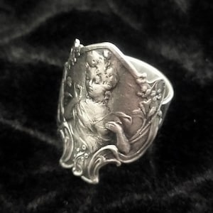 Könnte beinhalten: Ein silberner Ring mit Jugendstil-Design. Der Ring zeigt ein detailliertes Relief einer Frau, umgeben von floralen Elementen. Der Ring hat ein breites Band und ein verziertes Design, ideal, um einen Hauch von Vintage-Eleganz zu verleihen.