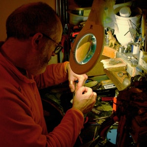 May include: A person working at a workbench, illuminated by a magnifying lamp. The person is wearing an orange long-sleeved shirt and glasses, focused on detailed work with small tools. The workspace is cluttered with tools and materials.