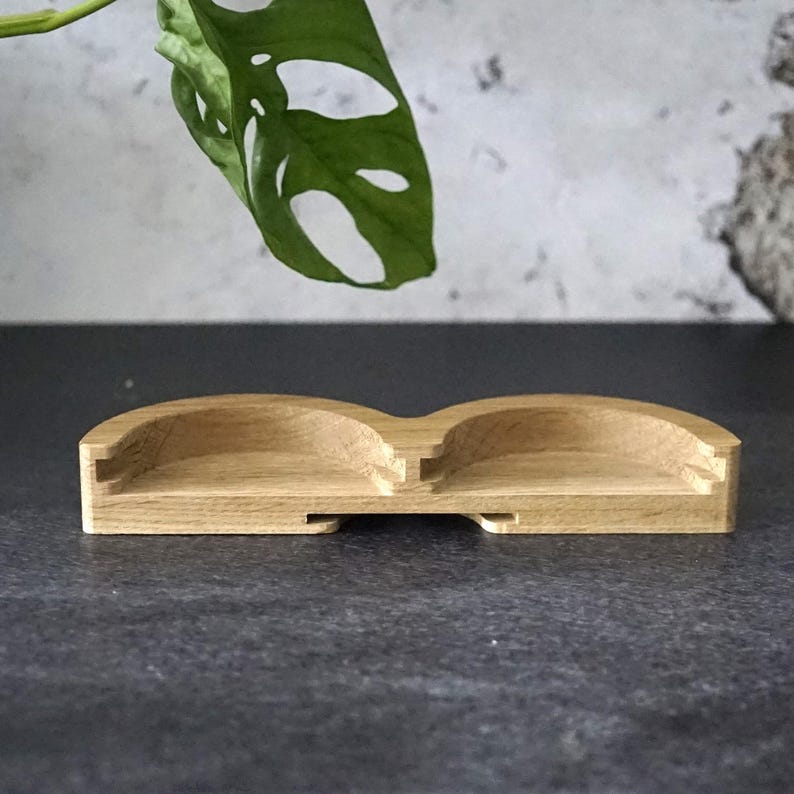 May include: A small, light-colored wooden tray with two curved compartments. The tray has a rectangular base and is made of natural wood. The design is simple and minimalist, suitable for holding small items. A green leaf is visible in the background.