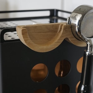 Op de afbeelding: Een close-up van een houten portafilterhouder bevestigd aan een zwarte espressomachine. De portafilterhouder is gebogen en heeft een uitsparing voor de portafilterhendel. De portafilter is gemaakt van roestvrij staal en heeft een gaasmand.