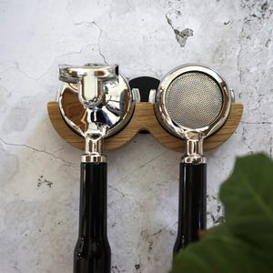 May include: Two espresso machine portafilters with shiny chrome heads and black handles are mounted on a wooden holder. The holder is made of light-colored wood and is mounted on a textured, light-colored wall.