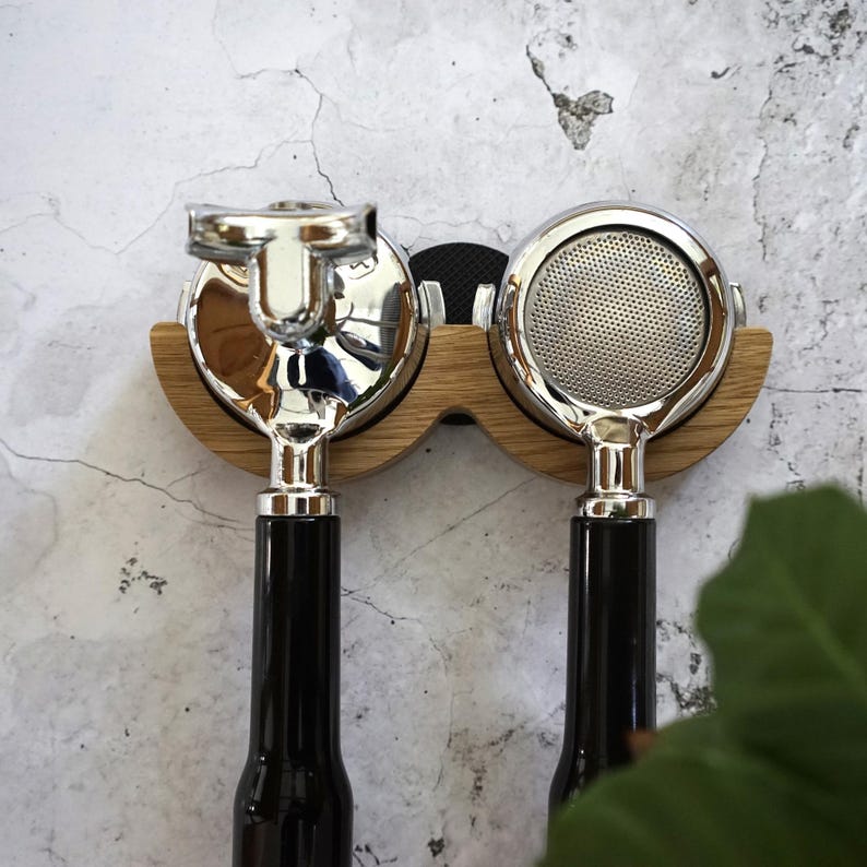 May include: Two espresso machine portafilters with shiny chrome heads and black handles are mounted on a wooden holder. The holder is made of light-colored wood and is mounted on a textured, light-colored wall.