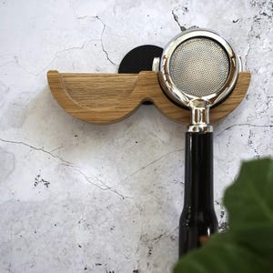 May include: A wooden espresso machine portafilter holder with a curved design. The holder is made of light-colored wood and features a black circular pad. A chrome portafilter with a black handle is inserted into the holder.