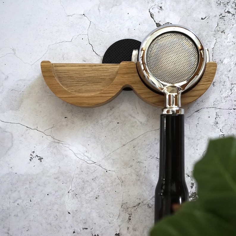 May include: A wooden espresso machine portafilter holder with a curved design. The holder is made of light-colored wood and features a black circular pad. A chrome portafilter with a black handle is inserted into the holder.