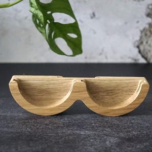 May include: A light brown wooden eyeglasses holder with two curved compartments. The holder is made of wood and has a natural grain pattern. The holder is designed to hold eyeglasses and is a stylish and functional accessory.
