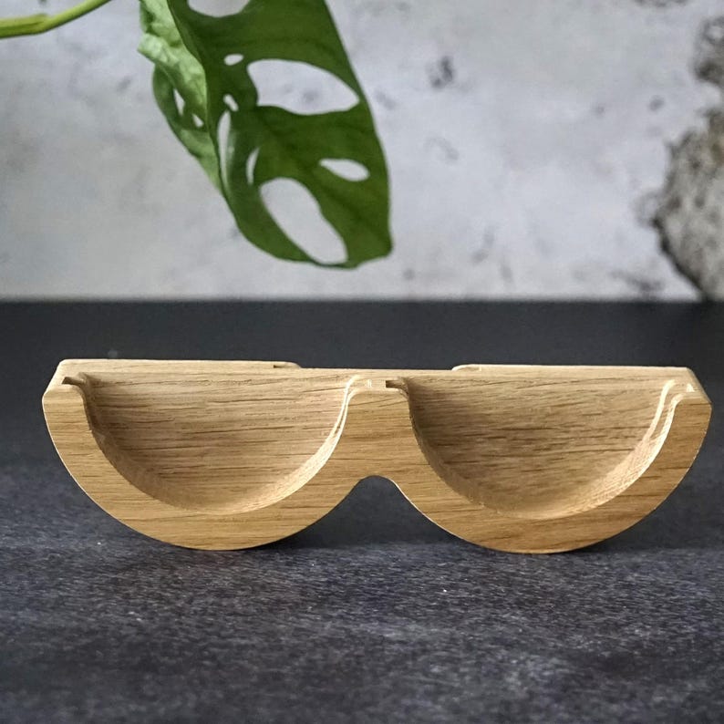 May include: A light brown wooden eyeglasses holder with two curved compartments. The holder is made of wood and has a natural grain pattern. The holder is designed to hold eyeglasses and is a stylish and functional accessory.
