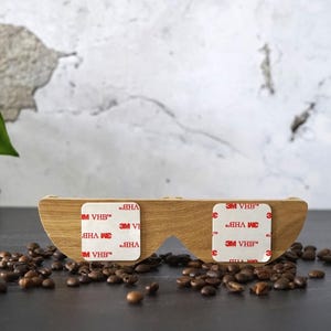 May include: A wooden, curved object with two cut-out sections, featuring three white adhesive pads with "3M VHB" text. The object is surrounded by coffee beans. The background is a textured, light-colored wall.