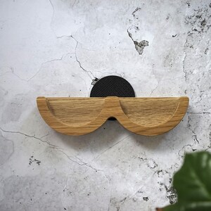 May include: A wooden wall shelf with a unique, curved design resembling a pair of glasses. The shelf is made of light-colored wood and is mounted on a black, circular backing. The shelf is mounted on a textured, light-colored wall.