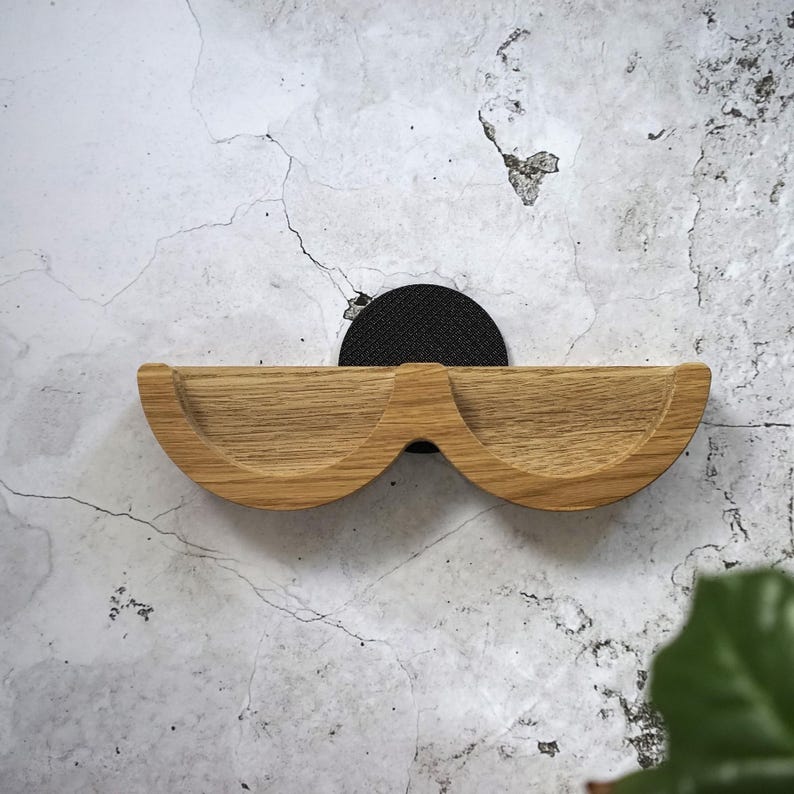 May include: A wooden wall shelf with a unique, curved design resembling a pair of glasses. The shelf is made of light-colored wood and is mounted on a black, circular backing. The shelf is mounted on a textured, light-colored wall.