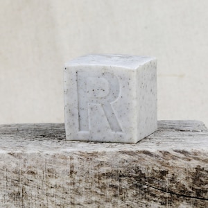 以下が含まれることがあります： 前面に「R」の文字が浮き彫りになった白い石鹸。石鹸は斑点模様になっている。