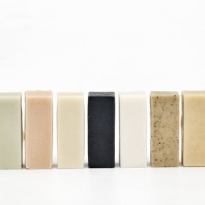 May include: A row of seven rectangular bars of soap in various colors, including green, beige, black, and white. The soaps are arranged side by side against a white background, showcasing their different textures and hues. The soaps are a natural product.
