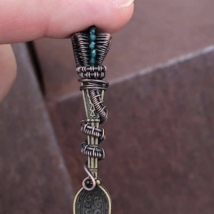 Puede incluir: Un colgante de cuchara envuelto en alambre de cobre con un acento de piedra preciosa verde. La cuchara tiene un diseño adornado y está unida a un bucle de alambre de cobre.