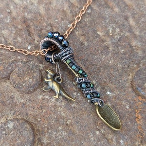 Puede incluir: Un collar de cadena de cobre con un colgante hecho de alambre envuelto con cuentas negras y verdes. El colgante presenta un pequeño amuleto de dinosaurio y un amuleto en forma de cuchara con un diseño grabado.