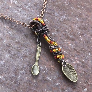 Puede incluir: Un collar de cadena de cobre con un colgante hecho de alambre envuelto con cuentas naranja, amarillo y negro. El colgante tiene dos cucharas pequeñas unidas a él.