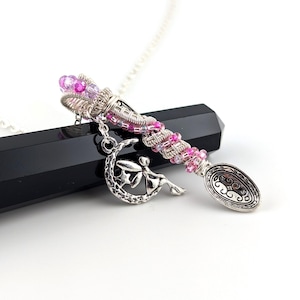 Puede incluir: Collar con colgante plateado con un dije en forma de cuchara adornado con cuentas rosas y moradas. Un dije de hada cuelga de la cuchara, descansando sobre una media luna. La cuchara tiene un diseño ornamentado.