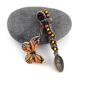May include: A handcrafted pendant featuring a butterfly charm and a spoon-shaped element. The pendant has a copper wire design with orange, yellow, and black beads. The butterfly has orange, black, and yellow wings. The pendant is approximately 3 inches long.