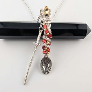 Può includere: Una collana con pendente in argento con un ciondolo a forma di spada in miniatura e un ciondolo a forma di cucchiaio con un design ornato. Il pendente è decorato con perline rosse e dorate e la catena è d'argento.