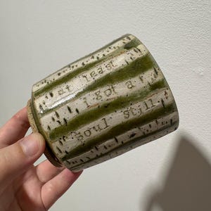 May include: A ceramic cup with a speckled off-white and olive green striped glaze. The exterior features the stamped text "at least I got a soul still" in a dark, recessed font. The base is unglazed, showing a natural tan clay texture.