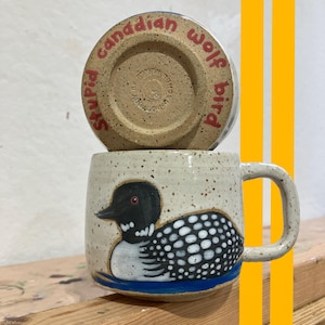Puede incluir: Una taza de cerámica moteada con un diseño de somorgujo pintado a mano. El ave tiene la cabeza negra, el cuerpo con estampado blanco y negro y un ojo rojo. La base de la taza tiene el texto "Stupid Canadian Wolf Bird" en rojo.