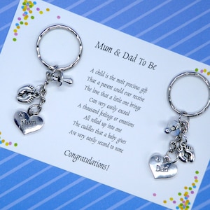 May include: Two silver keychains with heart-shaped charms that say "Mum" and "Dad" on them. The keychains also have charms of a baby's footprint, a dummy, and a bell. The keychains are on a blue and white striped card with the text "Mum & Dad To Be" and a poem about the love of a child.