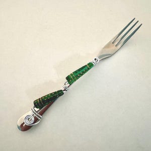 May include: A silver fork with a green and gold beaded handle. The beads are wrapped around the fork's handle with wire.