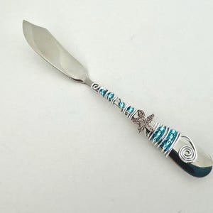 May include: A stainless steel butter knife with a decorative handle. The handle is wrapped in silver wire and adorned with turquoise beads and a starfish charm. The knife has a curved blade and a rounded end.