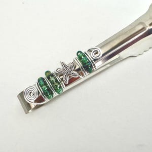 May include: Silver-toned serving tongs with decorative accents. Features green glass beads, silver wire swirls, and a starfish charm. The tongs are designed for serving food.