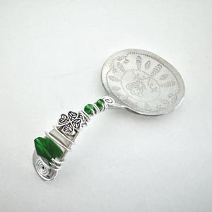 May include: A decorative silver spoon with a detailed floral design on the bowl. The handle is adorned with green beads, a green crystal, and a silver shamrock charm. The handle is wrapped in silver wire.