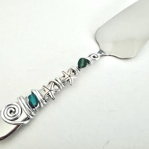 May include: Silver cake server with a decorative handle. The handle features silver wire wrapping, turquoise beads, small starfish charms, and dark green beads. The server has a wide, flat blade.