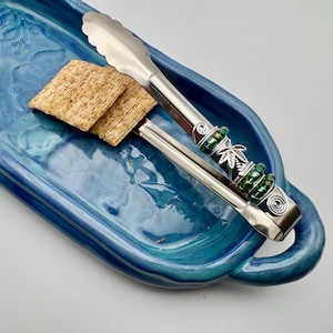 May include: A pair of silver serving tongs with green beaded accents and a leaf design on a blue ceramic serving tray with a handle. The tongs are holding two crackers.