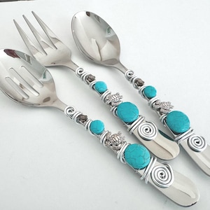 May include: A set of three stainless steel serving utensils: a slotted spoon, a fork, and a spoon. Each handle is adorned with turquoise-colored beads, silver wire, and small turtle charms. The utensils are arranged on a white surface.
