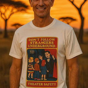 Könnte beinhalten: Weißes T-Shirt mit Retro-Grafik. Die Grafik enthält den Text "DON'T FOLLOW STRANGERS UNDERGROUND" und "THEATER SAFETY" sowie eine Illustration eines Mannes und Kindern. Der Hintergrund ist ein Sonnenuntergang.