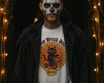 Puede incluir: Una persona con pintura facial de calavera lleva una camiseta blanca con un gráfico de Halloween. La camiseta presenta un gato negro montando en bicicleta con una cesta de calabaza, rodeado por el texto "Trick or Treat Patrol" y "Official".