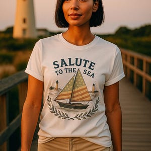 Op de afbeelding: Wit T-shirt met de tekst "SALUTE TO THE SEA" en een zeilbootafbeelding met een Amerikaans vlagontwerp op het zeil. De afbeelding toont een persoon die het shirt draagt, met een vuurtoren op de achtergrond.