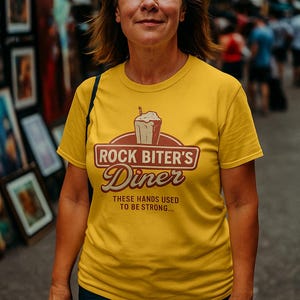 Pode incluir: Uma mulher vestindo uma camiseta amarela com o texto "ROCK BITER'S Diner" e um gráfico de milkshake. A camisa também tem a frase "THESE HANDS USED TO BE STRONG...". A mulher está ao ar livre.