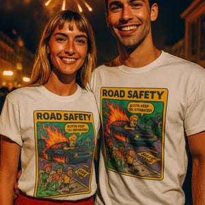 May include: Two people wearing white t-shirts with a colorful graphic promoting road safety. The graphic depicts a car accident with the text "ROAD SAFETY" and "GOTTA KEEP 'EM SEPARATED!" Fireworks are visible in the background.
