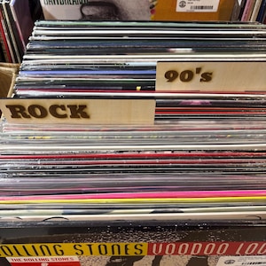 May include: A collection of vinyl records stored in a crate, featuring wooden dividers labelled ROCK and 90's. The front record is The Rolling Stones Voodoo Lounge, and a Jawbreaker record is visible in the back row with various other albums.