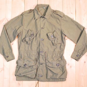 May include: An olive green military-style jacket with multiple pockets and button closures. The jacket has a collared neckline, long sleeves with buttoned cuffs, and a drawstring waist. It features four front pockets and epaulettes on the shoulders.