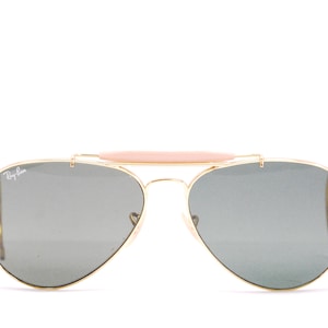 Pode incluir: &Oacute;culos de sol aviador dourados com ponte nasal marrom e lentes cinza. Os &oacute;culos t&ecirc;m o logotipo Ray-Ban na lente esquerda.