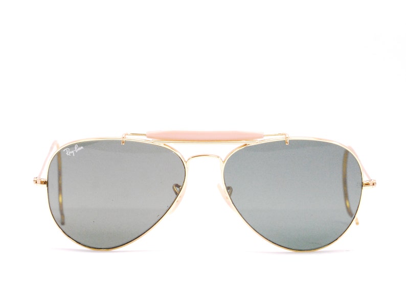 Pode incluir: &Oacute;culos de sol aviador dourados com ponte nasal marrom e lentes cinza. Os &oacute;culos t&ecirc;m o logotipo Ray-Ban na lente esquerda.