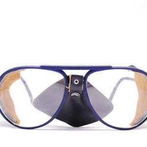 Puede incluir: Gafas de seguridad de estilo aviador azules con puente nasal de cuero marrón y lentes transparentes.