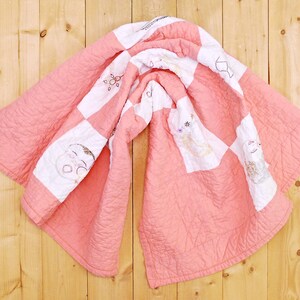 May include: A pink and white patchwork quilt with embroidered designs of children's faces and flowers. The quilt is folded and hanging on a wooden wall.