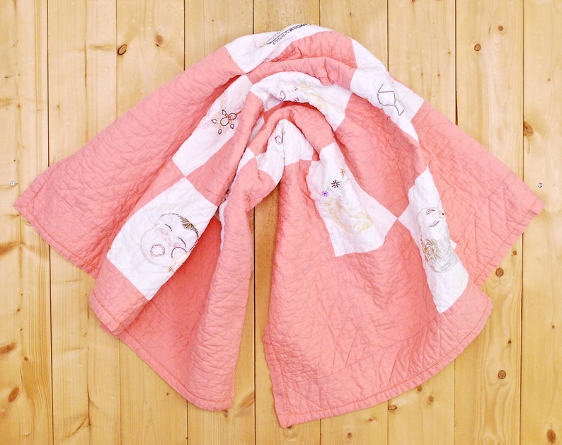 May include: A pink and white patchwork quilt with embroidered designs of children's faces and flowers. The quilt is folded and hanging on a wooden wall.