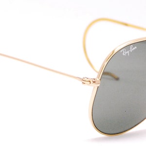 Pode incluir: &Oacute;culos de sol aviador dourados com lentes verdes e ponte nasal de couro marrom. Os &oacute;culos t&ecirc;m o logotipo Ray-Ban na lente direita.