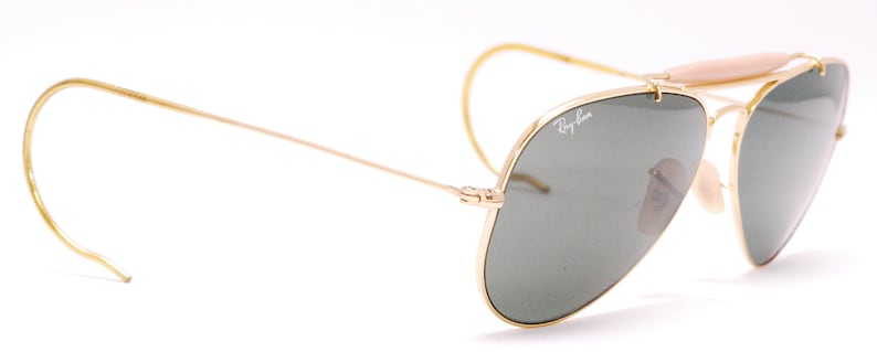 Pode incluir: &Oacute;culos de sol aviador dourados com lentes verdes e ponte nasal de couro marrom. Os &oacute;culos t&ecirc;m o logotipo Ray-Ban na lente direita.